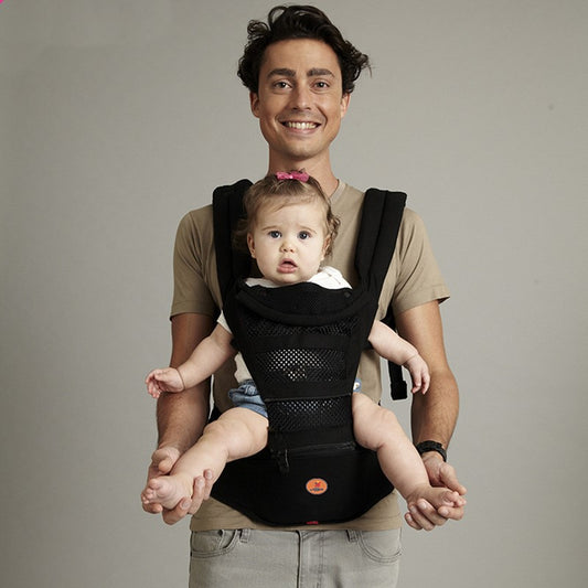 Smiling man carrying a baby in a black ergonomic baby carrier with a breathable mesh front and waist seat.