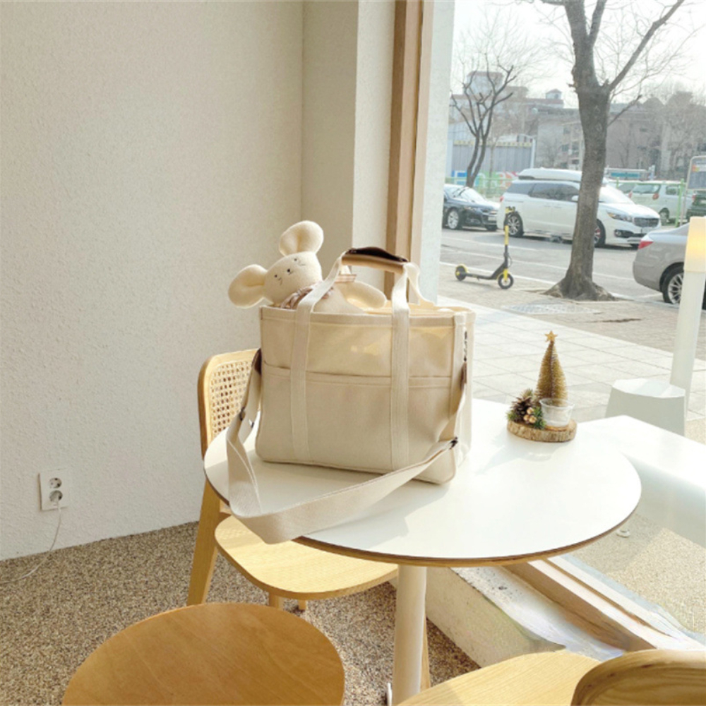 Stylish beige canvas diaper bag with shoulder strap, holding a plush toy, on a cafe table.