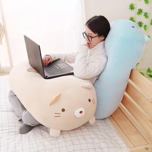 Woman using a laptop while leaning on a large, soft blue animal plush pillow and resting her arms on a beige animal plush.