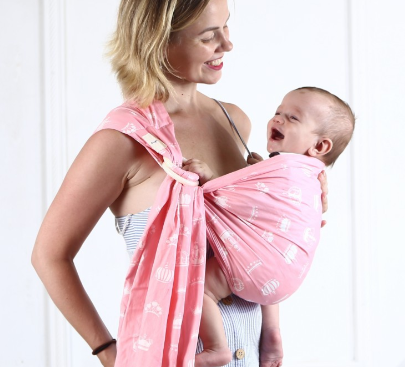 Woman smiling, carrying a happy baby in a pink cotton ring sling with white crown patterns.