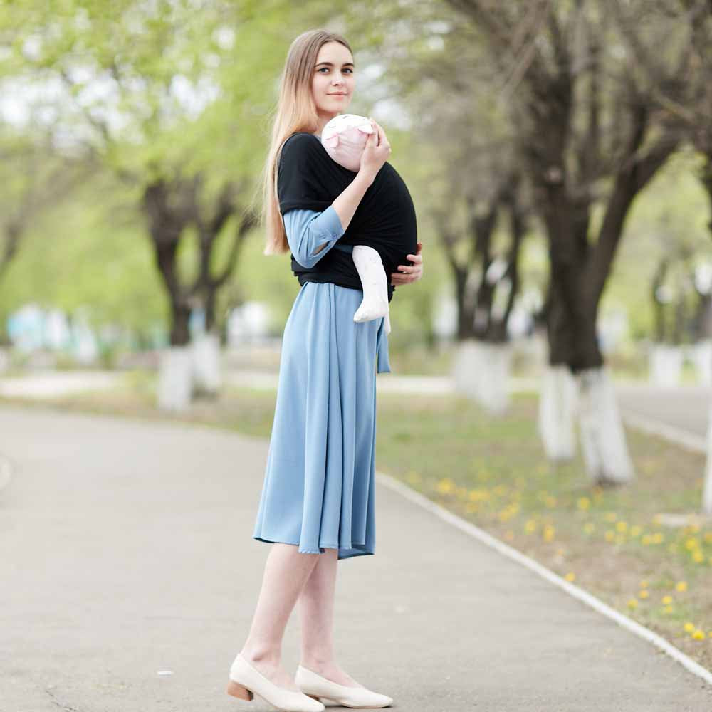 Woman in a blue dress holding a baby in a black cotton baby wrap carrier outdoors.
