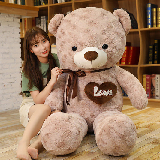Giant soft beige teddy bear with a brown bow and "Love" heart, hugged by a woman.
