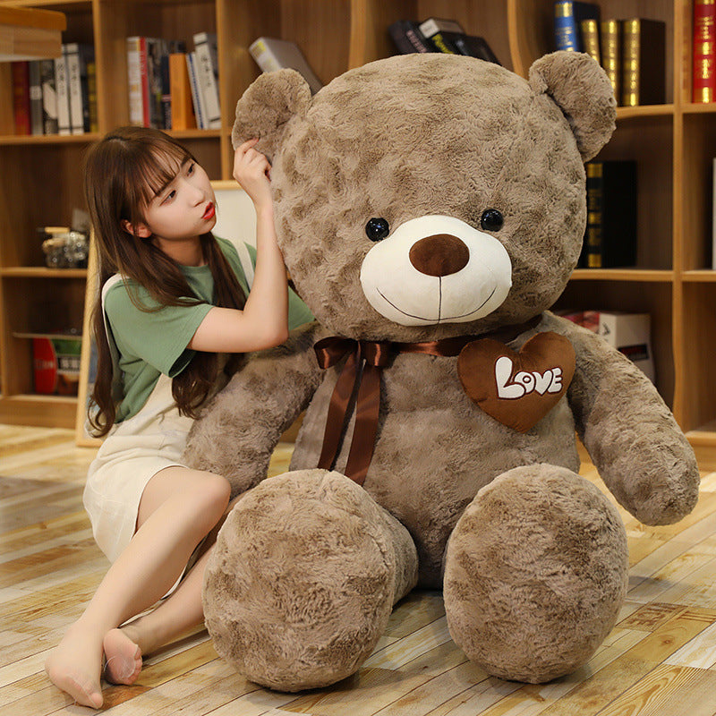 Large brown plush teddy bear with a "Love" heart and ribbon, next to a woman.