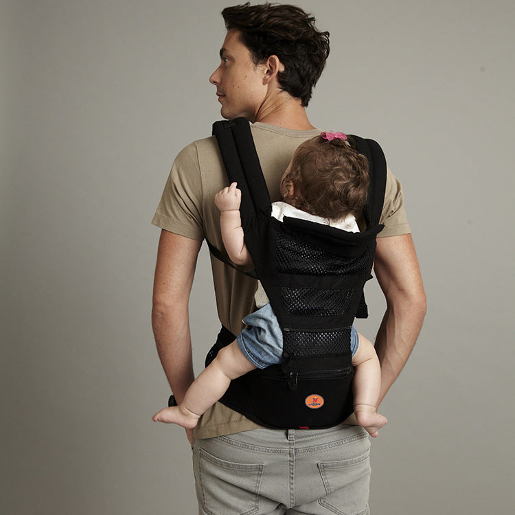 Man carrying baby in a black ergonomic baby carrier with waist seat and mesh panel.