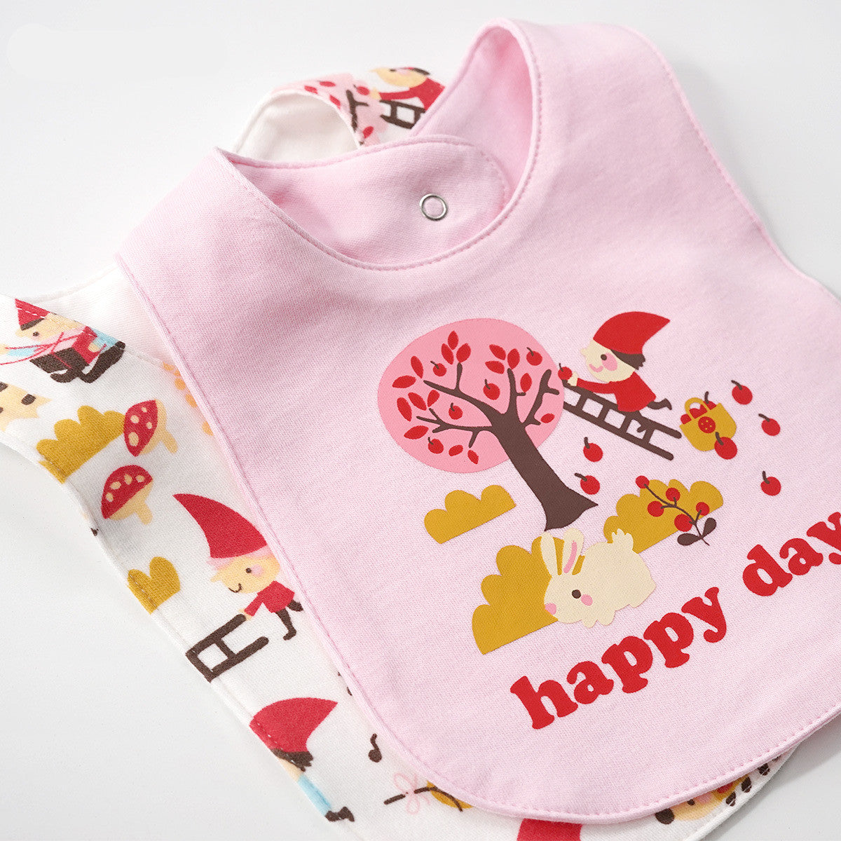 Pink cotton baby bib with "happy day" text and gnome picking apples, over a white patterned bib.