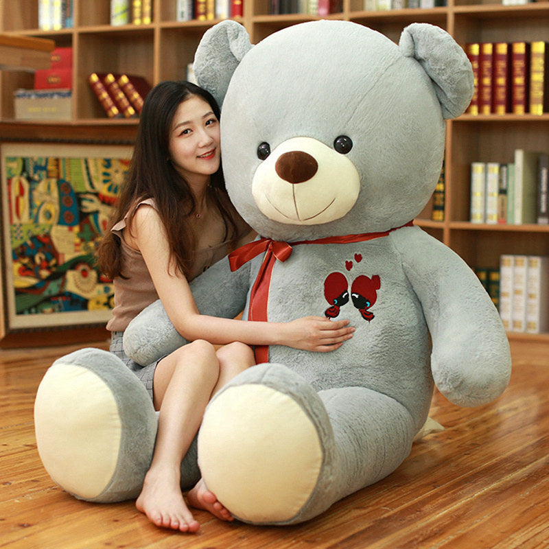 Large gray plush teddy bear with a red ribbon and heart embroidery, hugged by a woman.