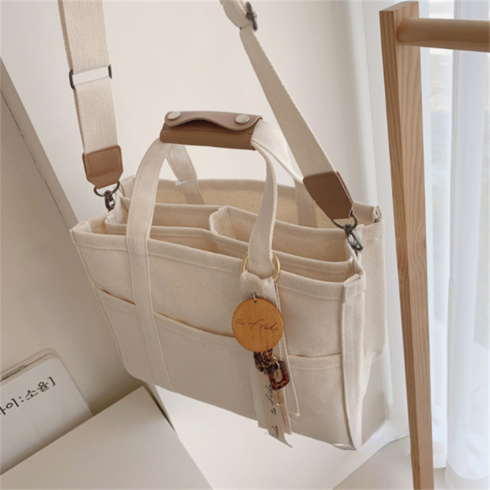 Cream canvas diaper bag with brown leather accents, multiple compartments, and a wooden charm.