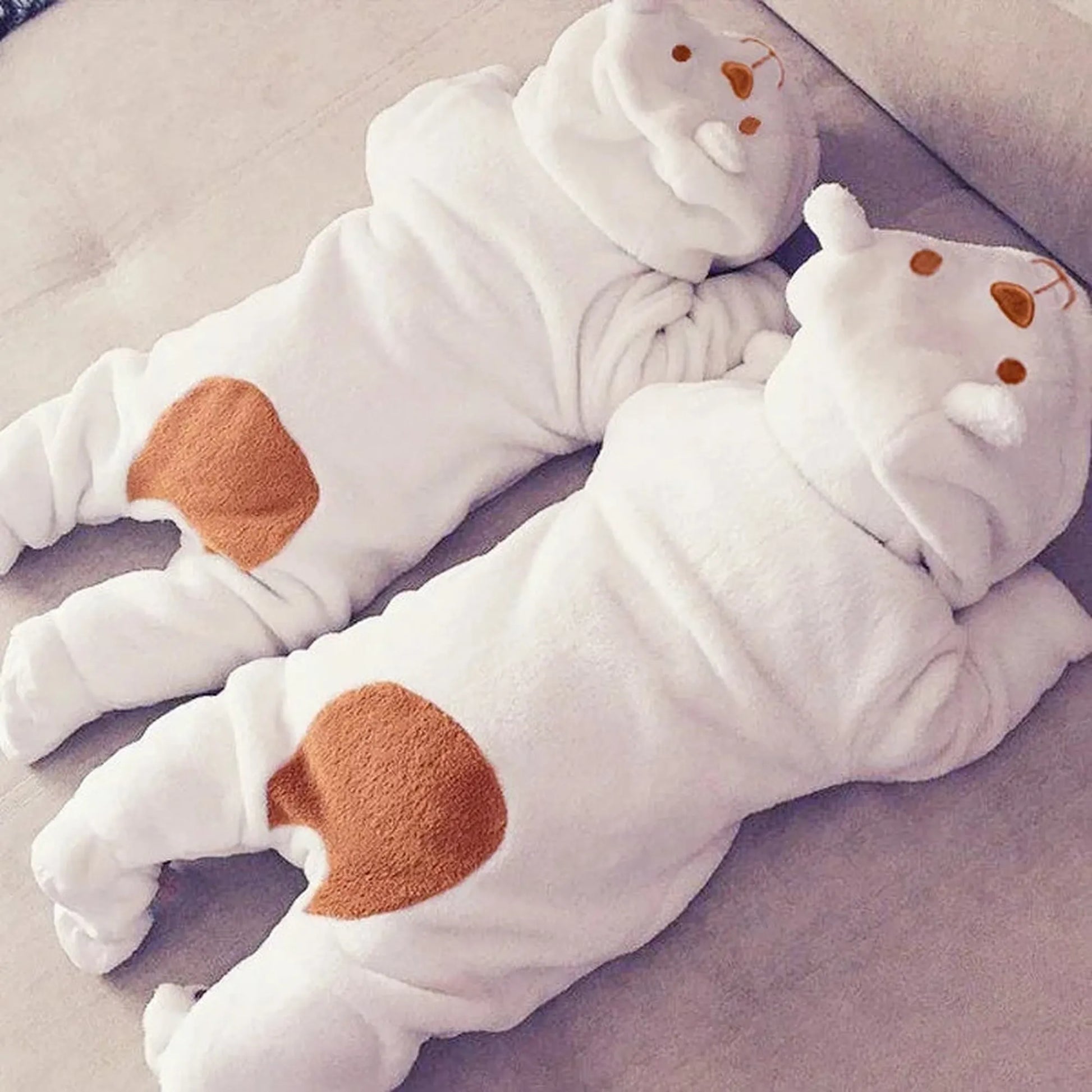 Two white plush baby bear hooded rompers with brown spots, lying on a light-colored surface.
