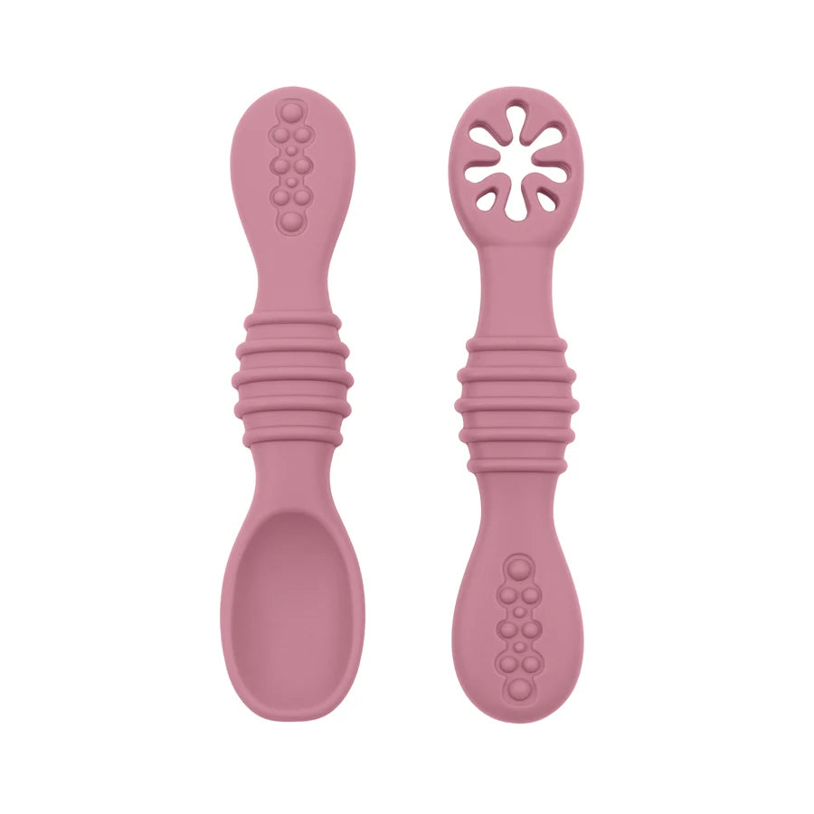 Pink silicone baby training spoons with textured handles and soft tips, one solid, one with a star-shaped opening.