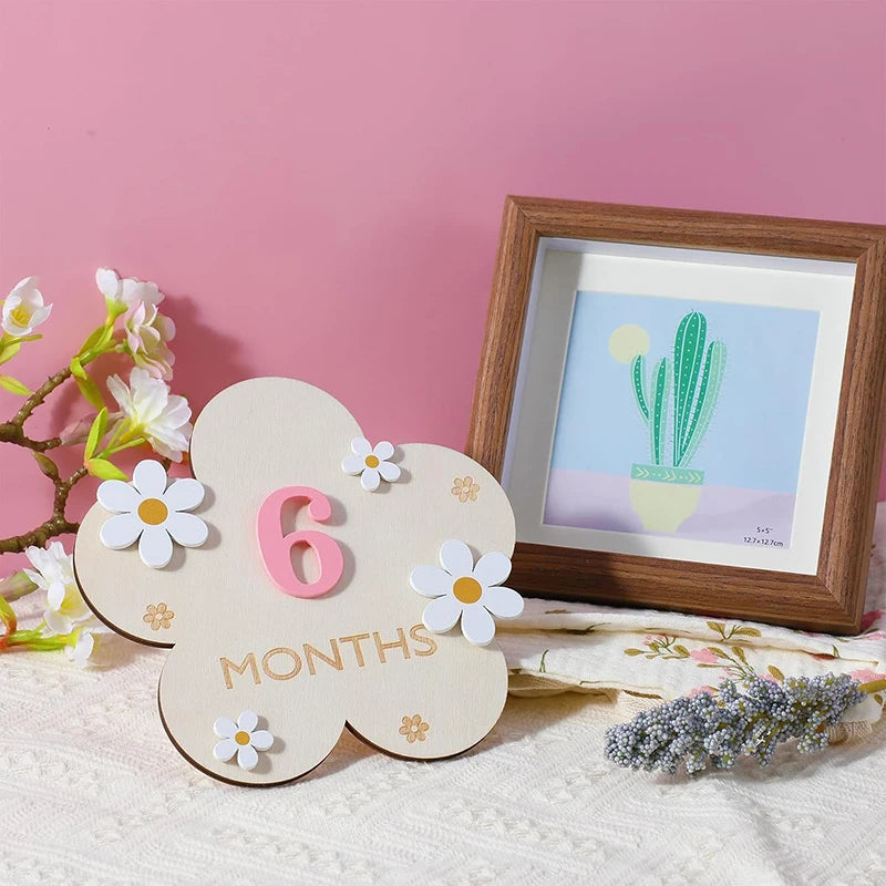 Wooden flower-shaped baby milestone card with "6 MONTHS" in pink and white, decorated with small white flowers.