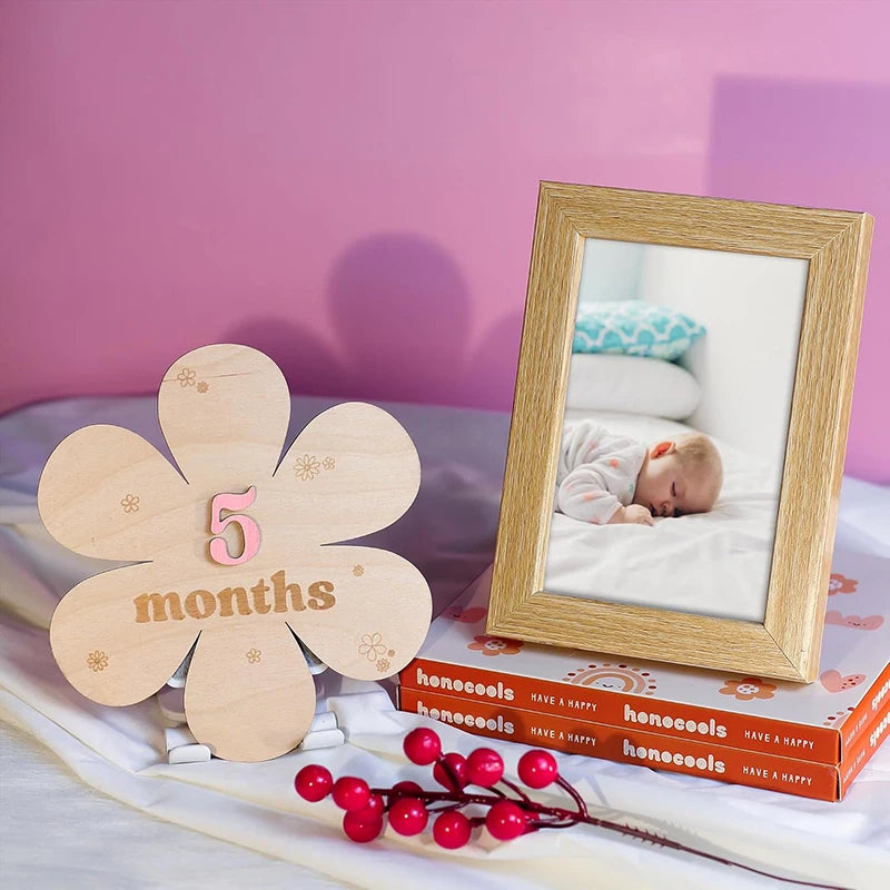 Wooden flower milestone card "5 months" next to a framed baby photo, with red berries and boxes.