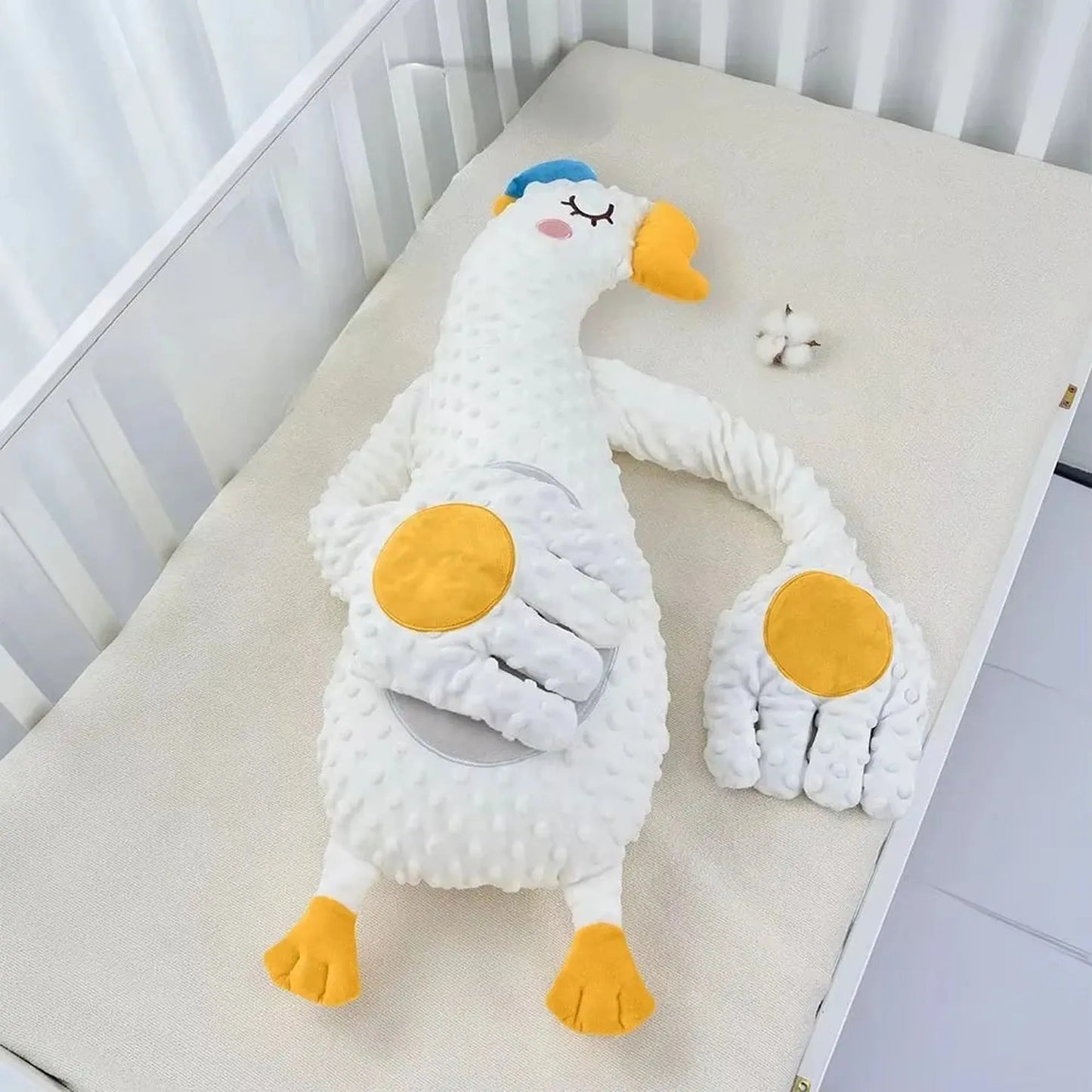 White plush goose baby soothing pillow with yellow accents and a detached hand, in a crib.