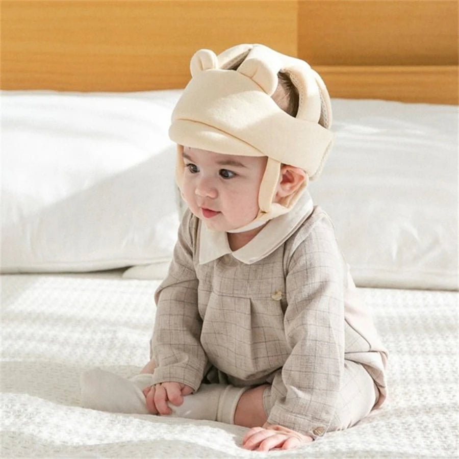 Baby wearing a beige, breathable anti-collision safety helmet with ear flaps, sitting on a bed.
