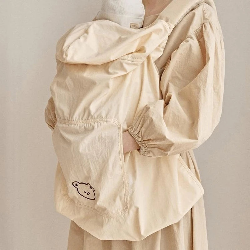Beige polyester baby carrier cover with a cute bear design and hood, worn over a baby.