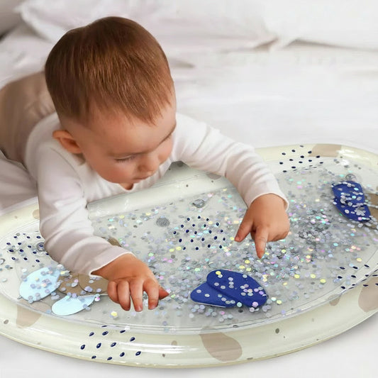 Baby on tummy time playing with an inflatable water mat filled with silver glitter and blue shapes.