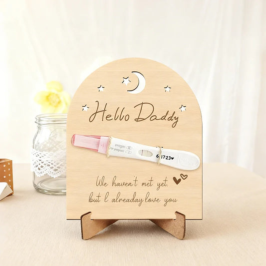 Arched wooden pregnancy announcement sign with "Hello Daddy" and "We haven't met yet, but I already love you" text, holding a positive pregnancy test.