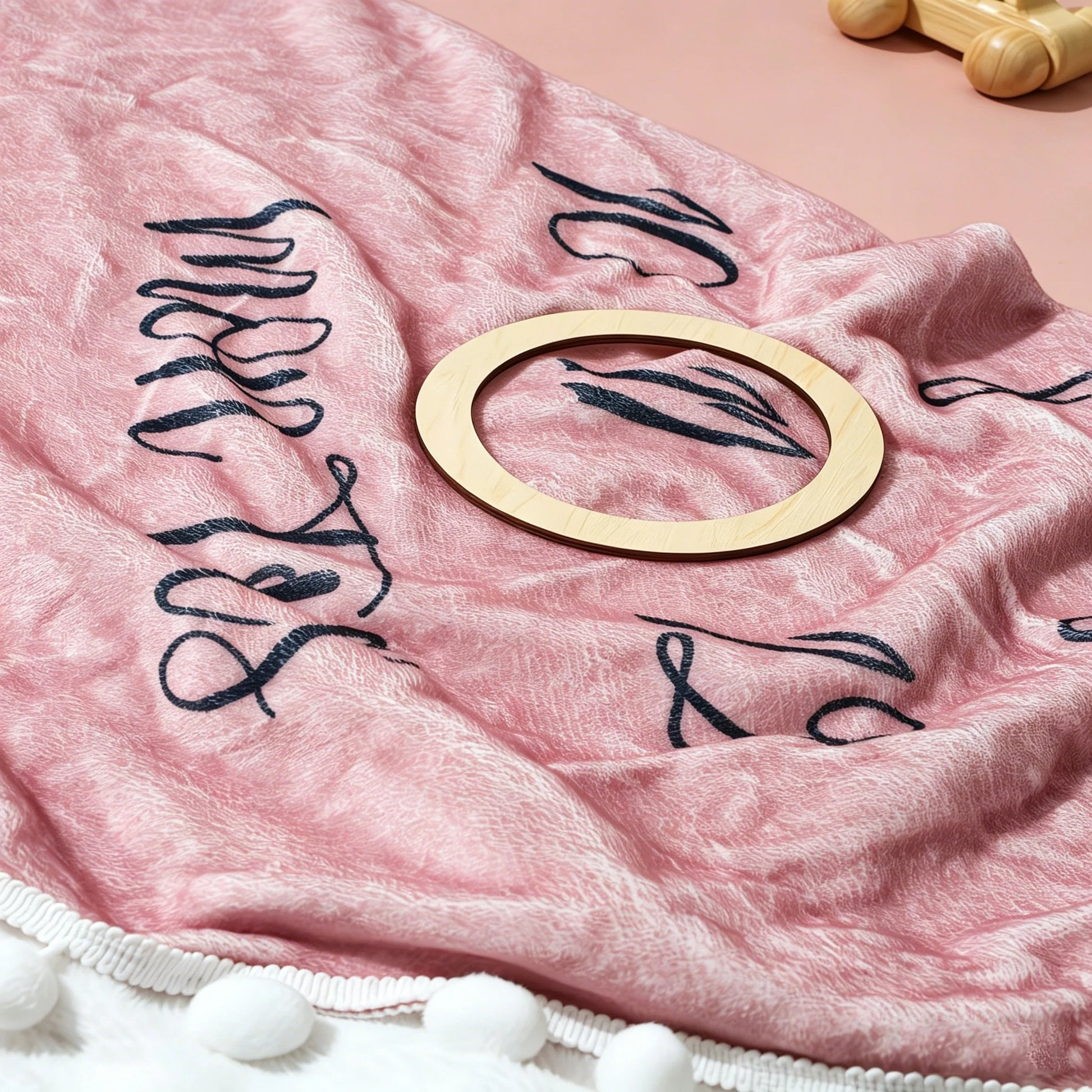 Pink flannel baby milestone blanket with black script, white pom-poms, and a wooden marker.