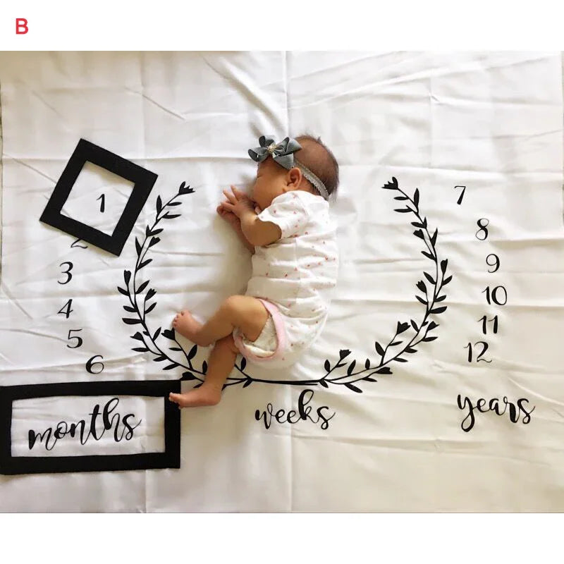 White polyester baby milestone blanket with black numbers, "months," and laurel wreath design, featuring a sleeping infant.
