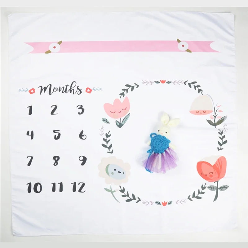 White polyester baby milestone blanket with floral design, "Months" text, numbers 1-12, and a bunny toy.