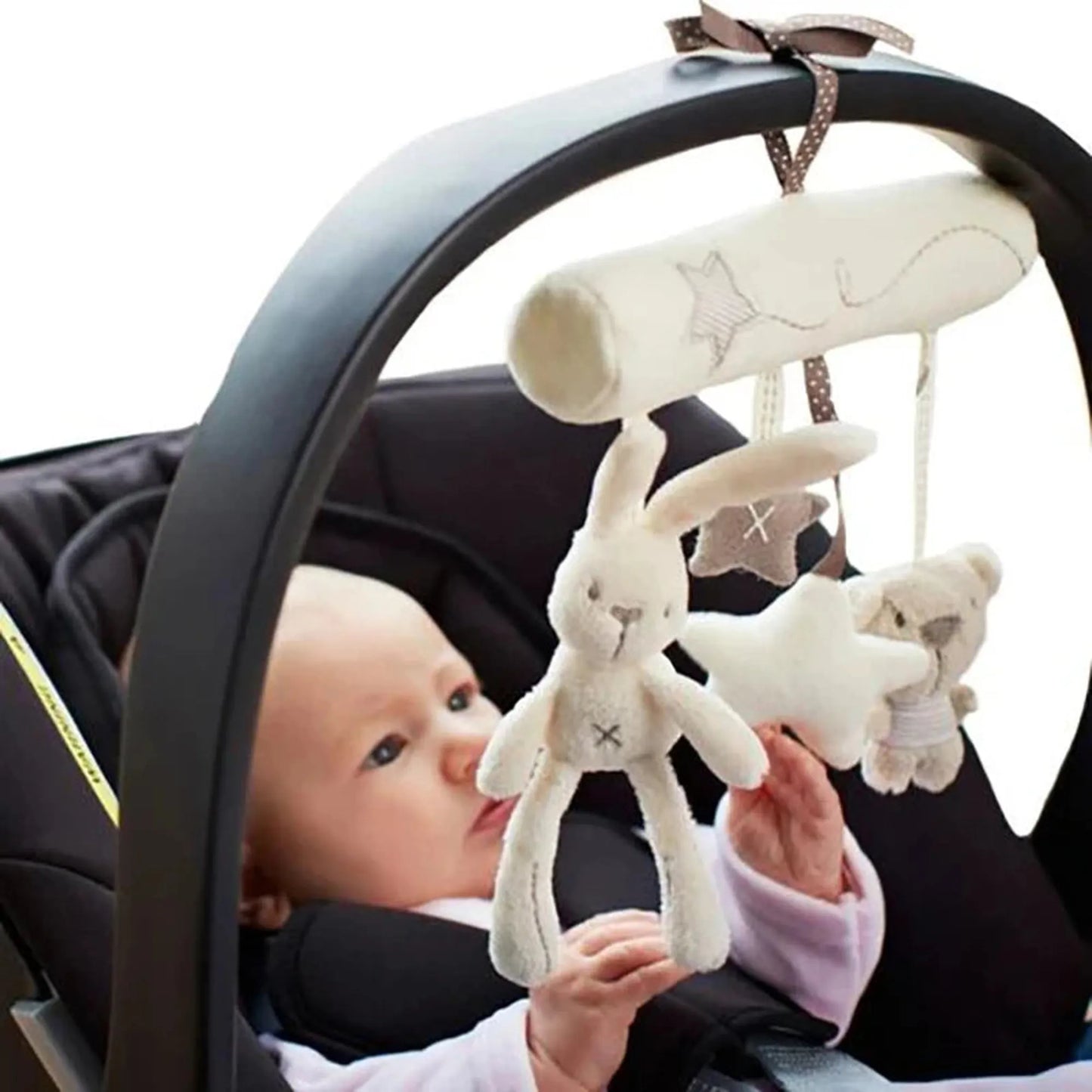 Plush bunny, star, and bear rattle toy hanging from a car seat bar, with a baby looking at it.