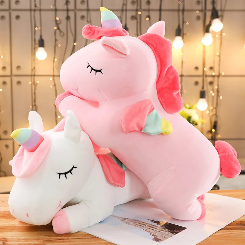Two soft plush unicorn toys, one pink and one white, with rainbow horns and closed eyes.
