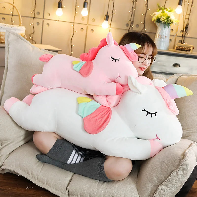 Woman cuddling a large white plush unicorn and a smaller pink unicorn with rainbow accents.