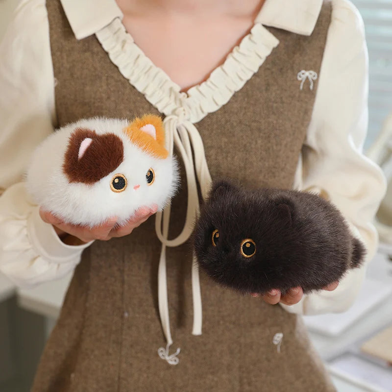 Two fluffy, round cat plush toys: one white, brown, and orange, the other dark brown, held by a person.
