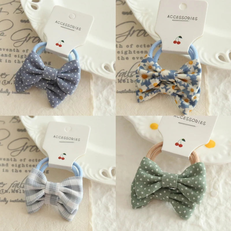 Four soft cotton hair ties for babies and girls, featuring bows in blue polka dot, floral, plaid, and green polka dot designs.