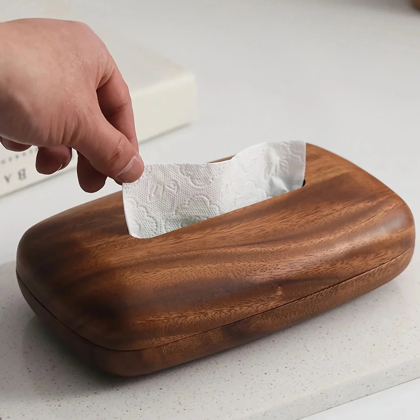 Hand pulling a white tissue from a rectangular vintage wooden tissue box with a smooth, dark brown finish.