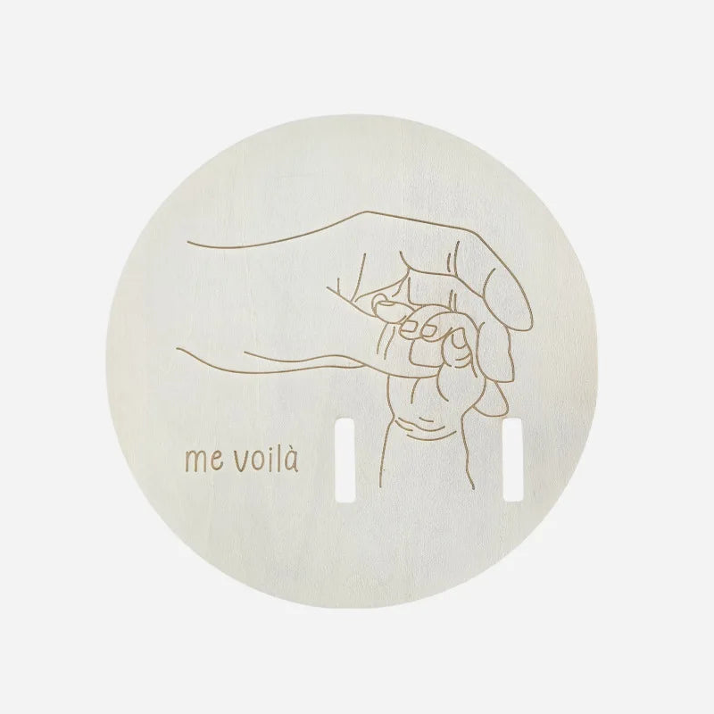 Round wooden birth keepsake board with engraved parent and baby hands, "me voilà" text, and slots for a hospital bracelet.