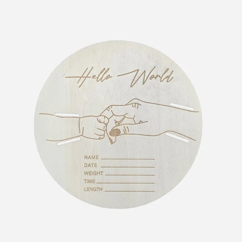Round wooden birth announcement sign with "Hello World" engraving, baby and adult hand illustration, and spaces for birth details.