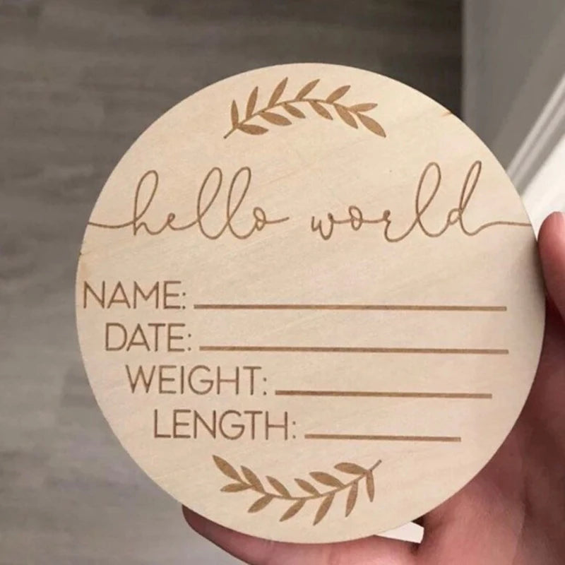 Round wooden "Hello World" birth announcement sign with engraved baby details and leaf accents.