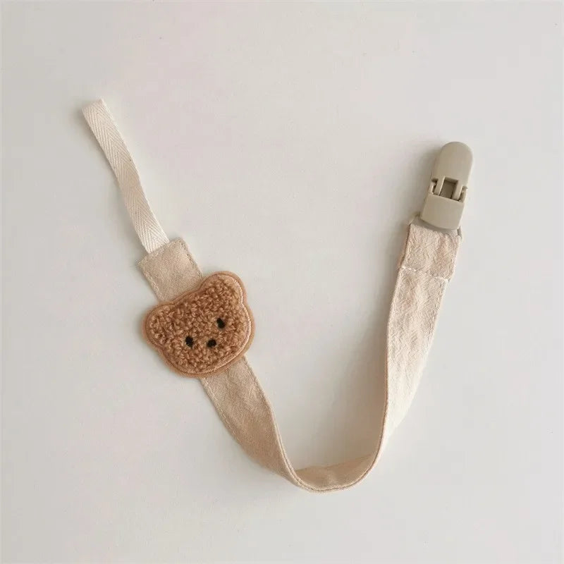Beige cotton pacifier clip with a cute embroidered teddy bear and a plastic clasp.
