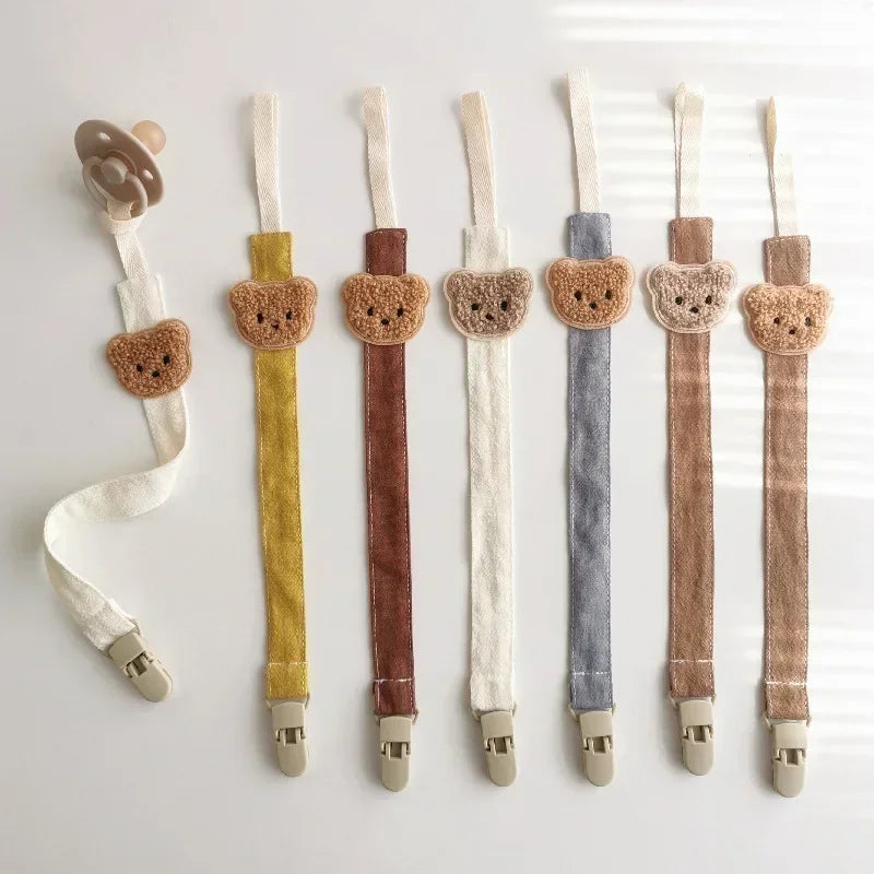 Seven cotton pacifier clips in various colors, each with a cute embroidered teddy bear and plastic clasp.