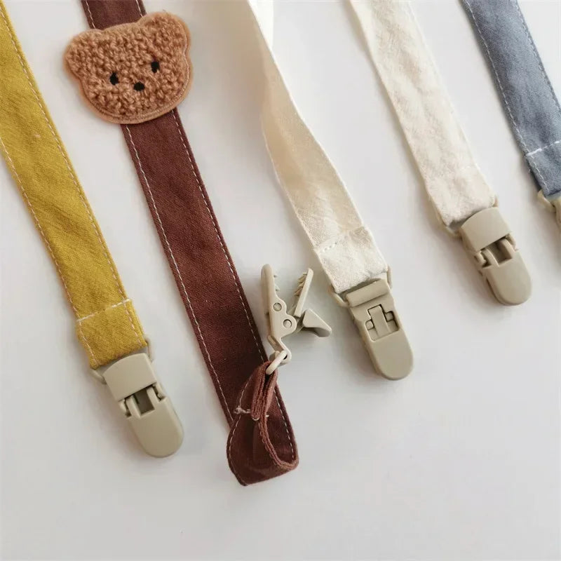 Cotton pacifier clips in yellow, brown with a teddy bear, white, and blue, featuring plastic clasps.