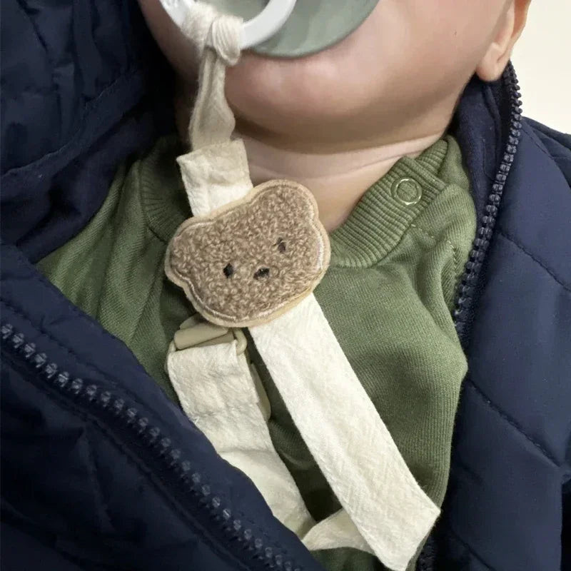 Baby wearing a cream cotton pacifier clip with a cute embroidered brown bear design on a green shirt.