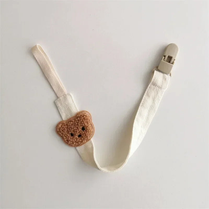 Cream cotton pacifier clip with embroidered brown teddy bear design and beige plastic clasp.