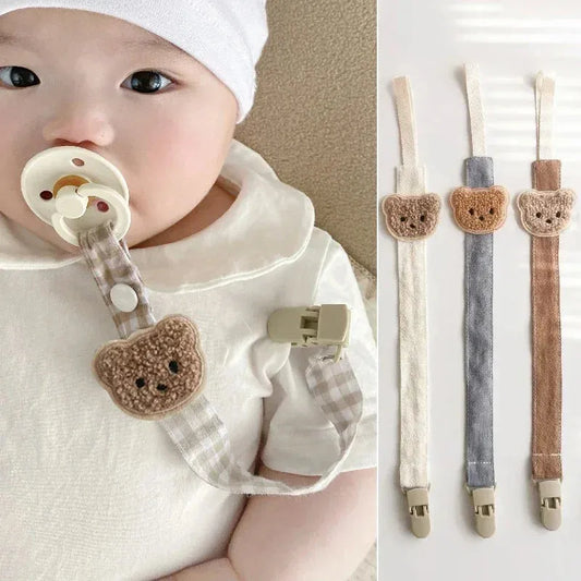Baby with a beige pacifier clip featuring a brown embroidered bear and checkered cotton strap. Three clips in white, gray, and brown are shown.