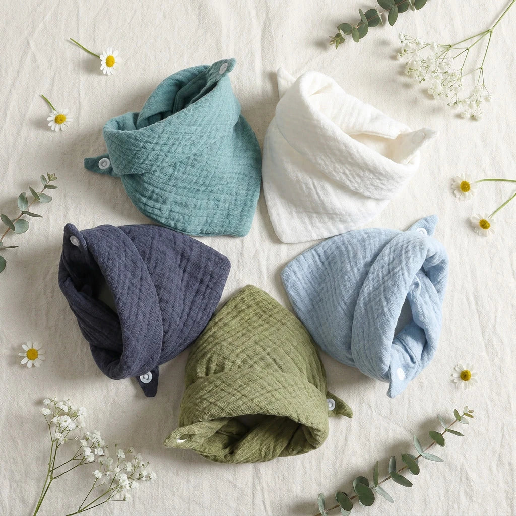 Five soft cotton baby bibs in white, blue, teal, olive, and navy, with snap closures, arranged on a light fabric.
