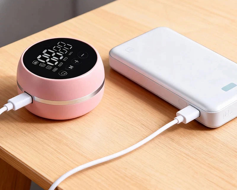 Pink wearable breast pump with digital display charging from a white power bank on a wooden table.