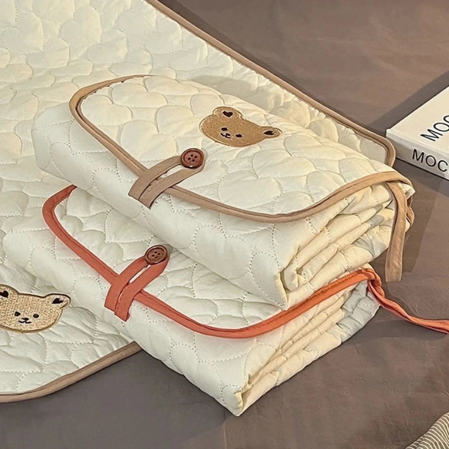 Two quilted, foldable, cream-colored cotton changing pads with embroidered bear designs and button closures.
