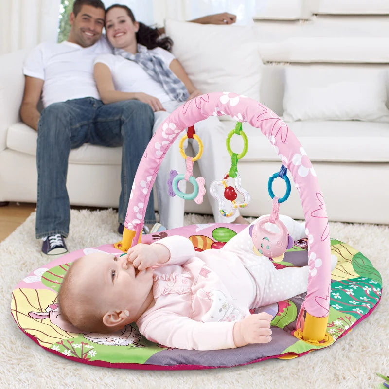 Baby on a pink floral play mat with hanging toys, parents relaxing in the background.