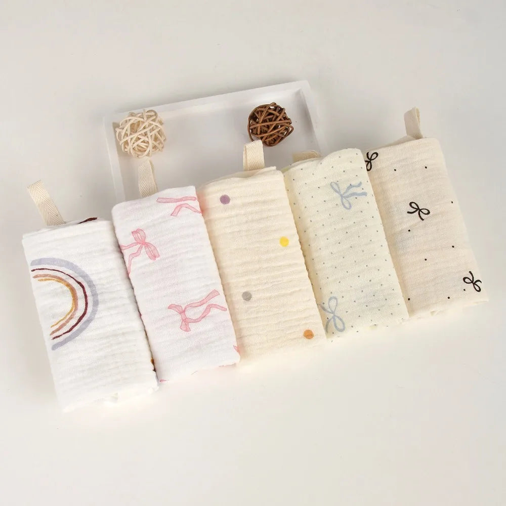 Soft white and cream multi-layered cotton gauze baby towels with various patterns like rainbows, bows, and dots.