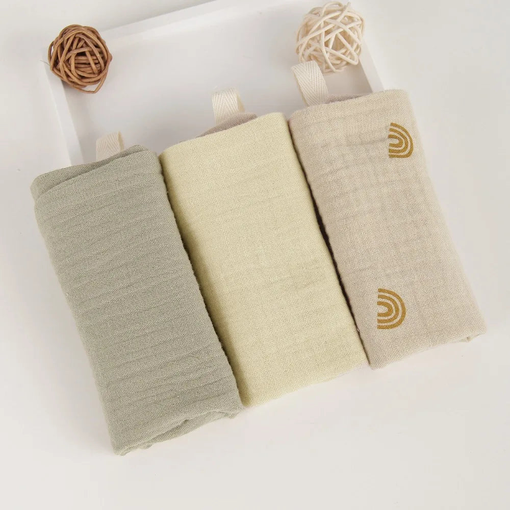 Three rolled baby gauze cotton towels in sage green, cream, and beige with rainbow embroidery.