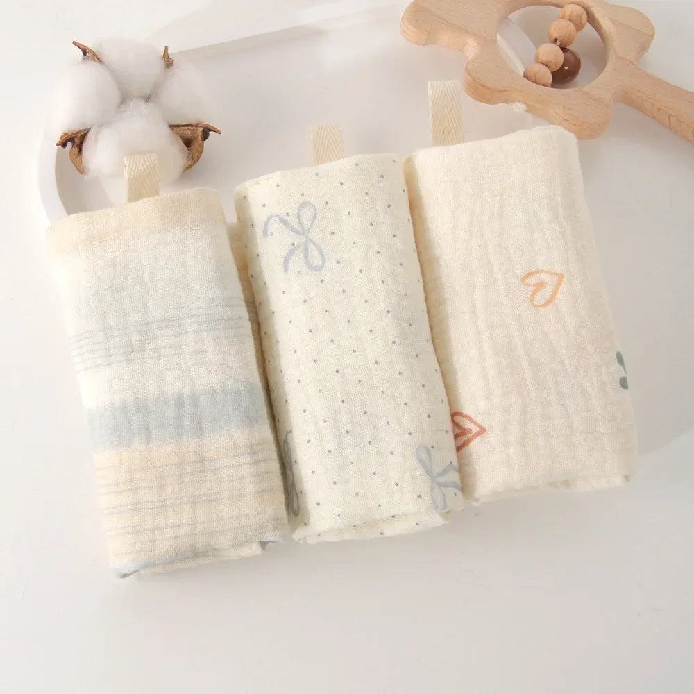 Three folded soft cotton gauze baby towels in cream, one striped, one with dots and bows, one with hearts.