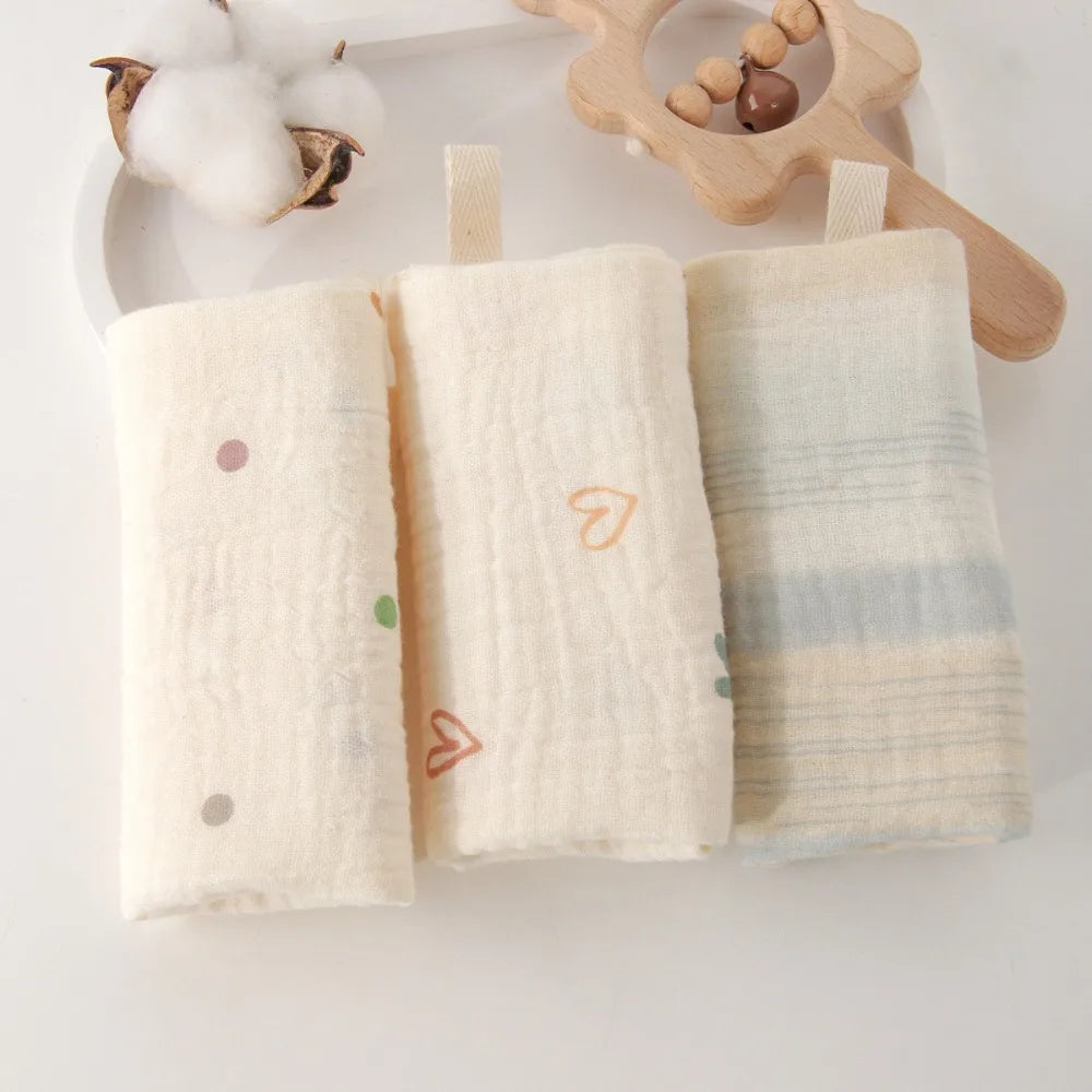 Three soft, natural cotton gauze baby towels: one with colorful dots, one with orange hearts, and one with blue stripes.