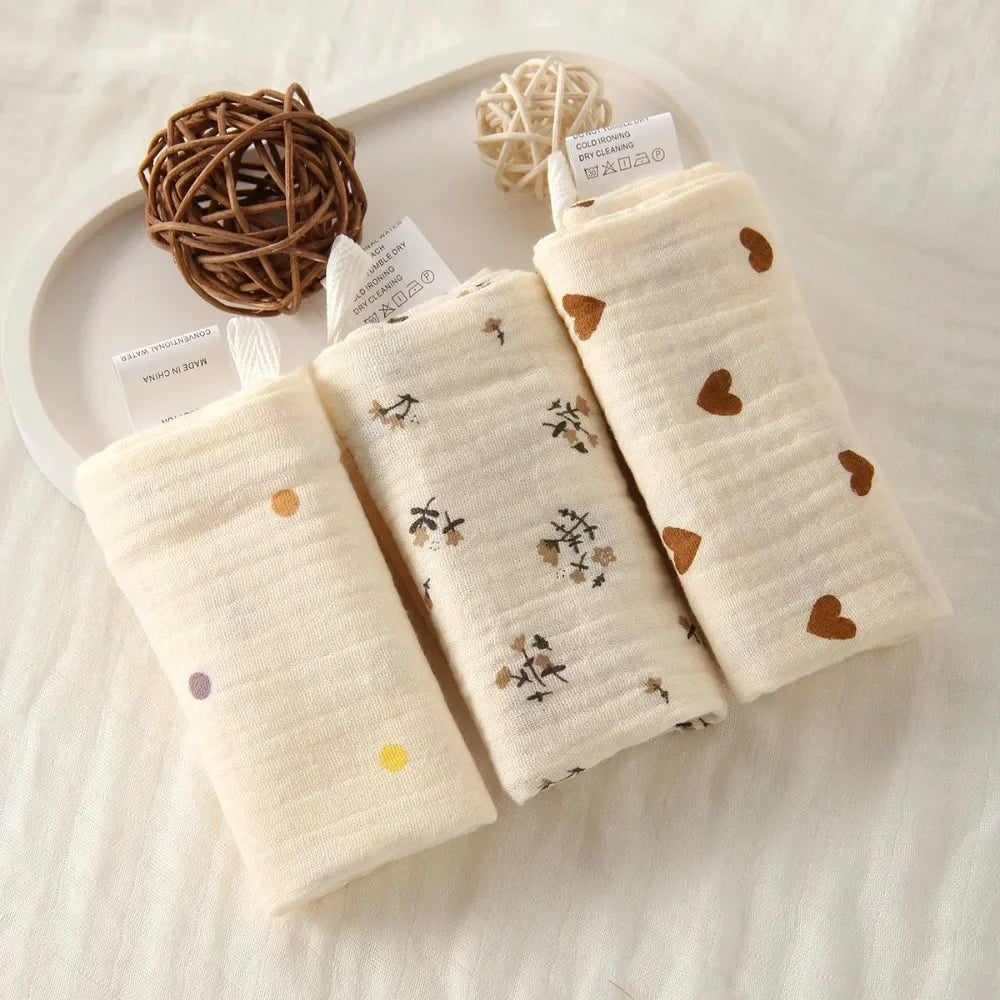 Three rolled natural cotton gauze baby towels with heart, floral, and dot patterns on a white tray.
