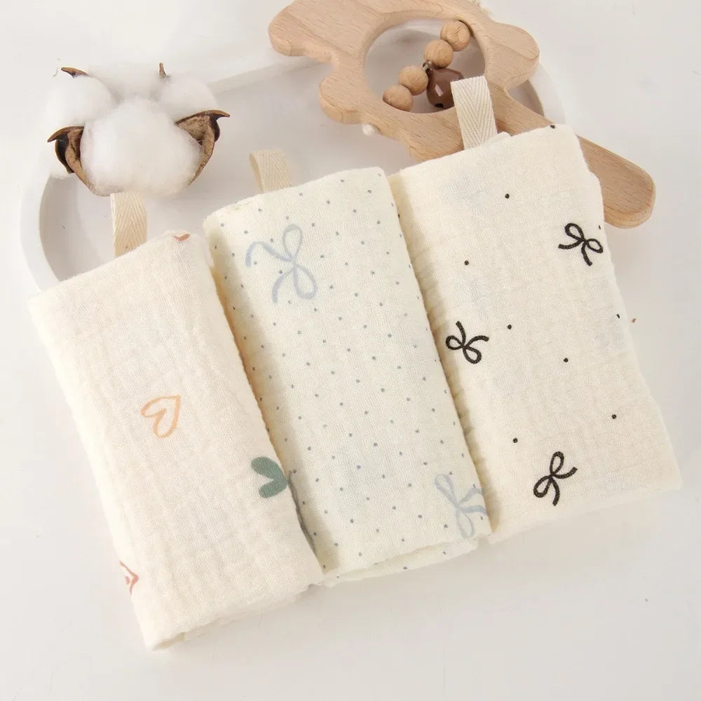 Three soft, natural cotton gauze baby towels with subtle patterns and hanging loops, beside a wooden rattle.