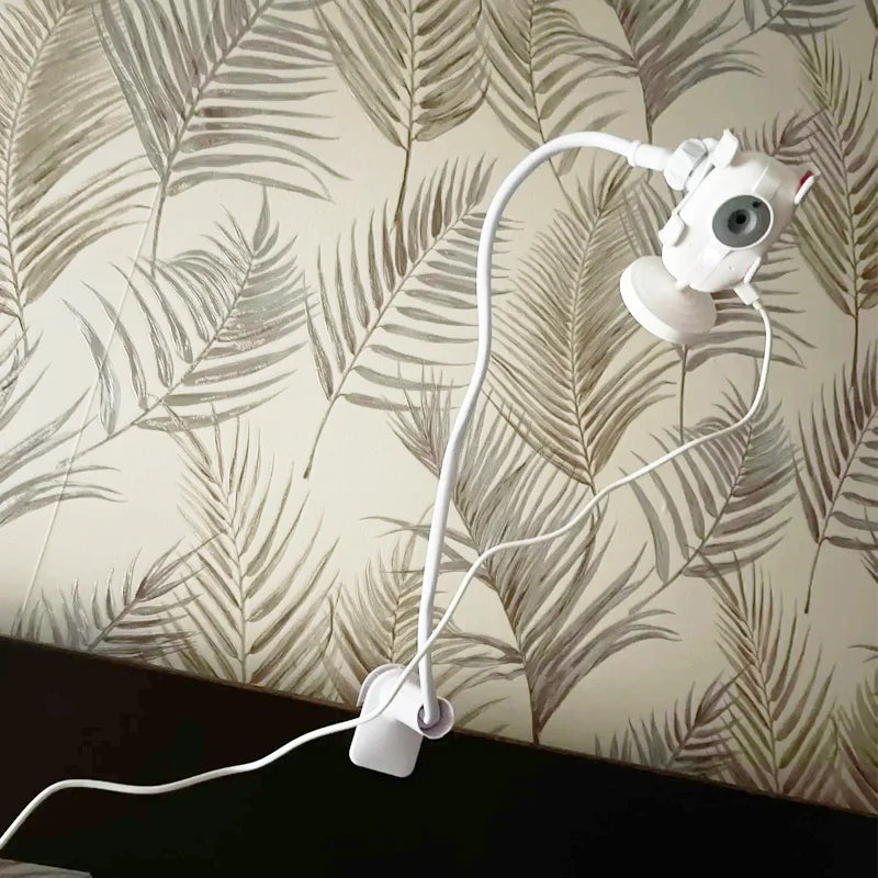 White flexible baby monitor holder with long adjustable arm and clamp, against palm leaf wallpaper.