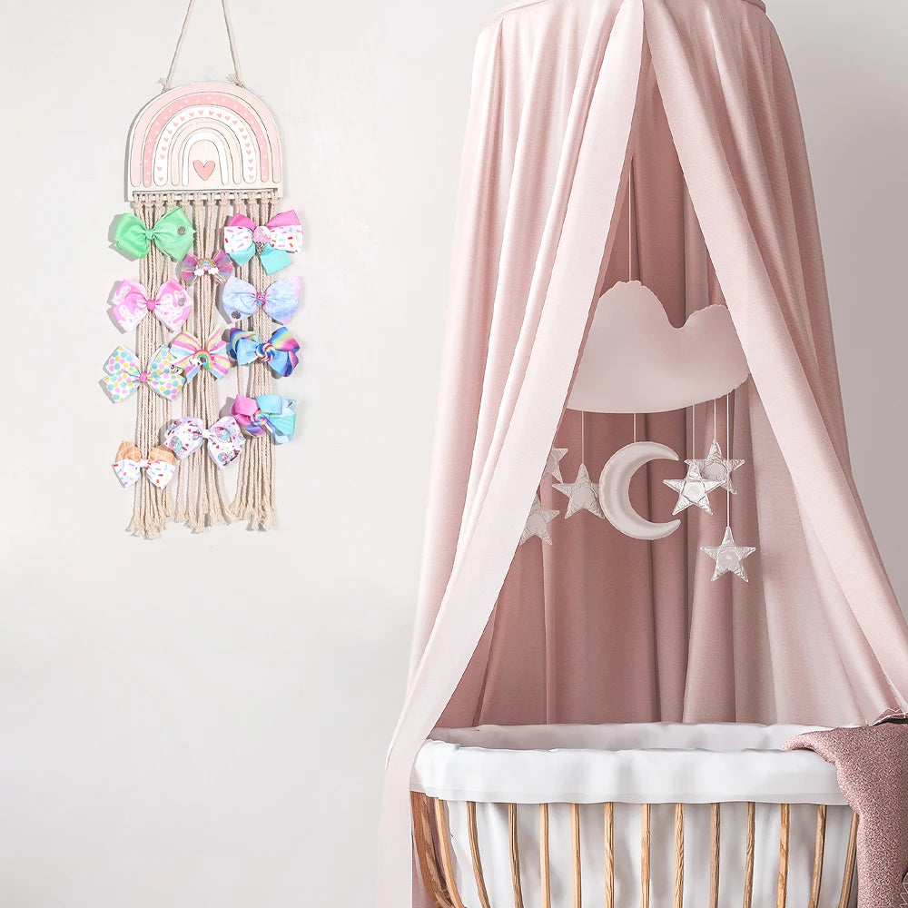 Pink rainbow macrame wall hanger for girls' hair bows, clips, and accessories, next to a baby crib.